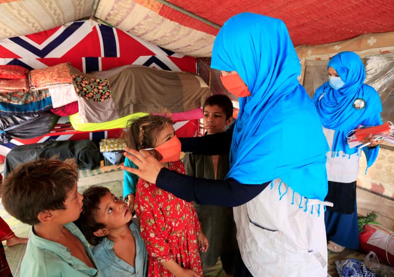 protect refugees: A UNICEF worker helps an internally displaced Afghan girl put on a protective mask at a makeshift camp in Jalalabad in late June during the COVID-19 pandemic. (CNS photo/Parwiz, Reuters)