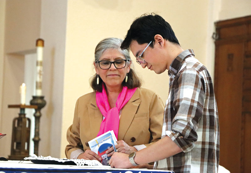 Sisolak, the son of a returned lay missioner, signs the Maryknoll Lay Missioners covenant pledging to serve in overseas mission while Executive Director Elvira Ramirez looks on. (Andrea Moreno-Díaz/U.S.)